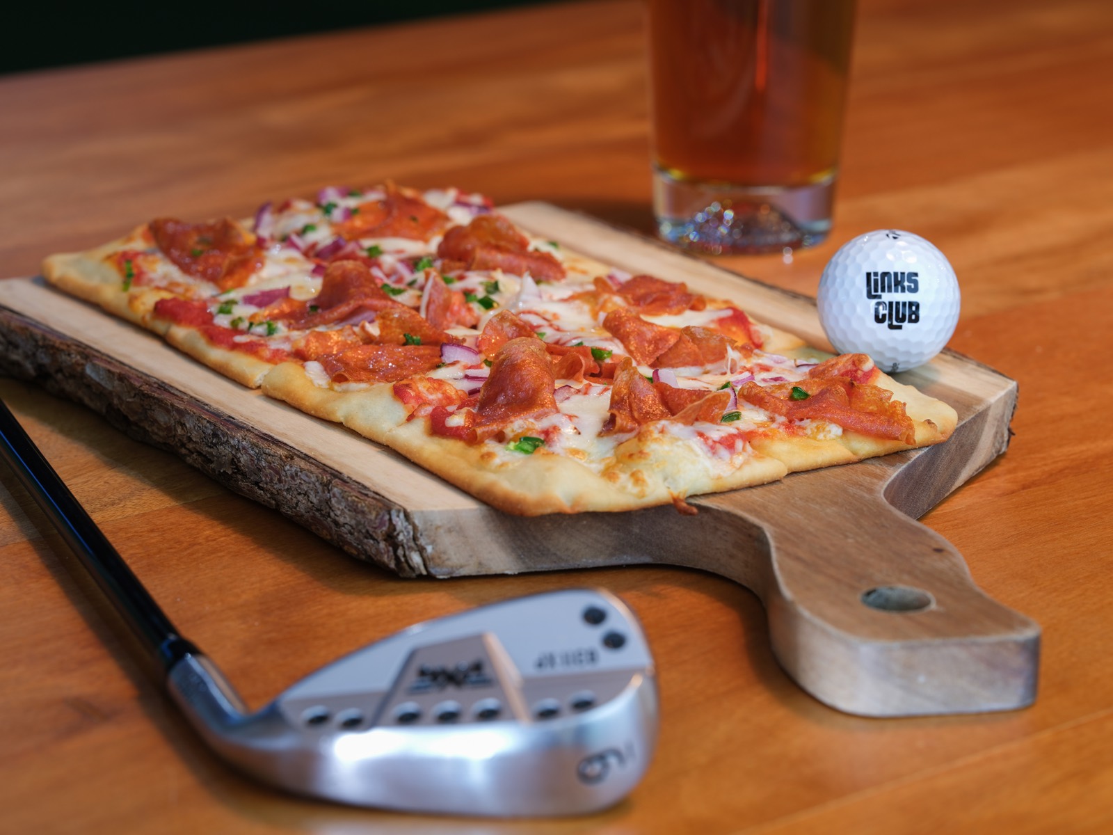 Pepperoni flatbread on a wood board beside a Links Club golf ball and craft beer